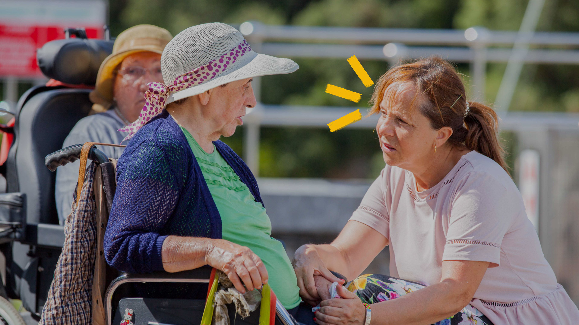 Dos mujeres mayores hablando, una de ellas en silla de ruedas. La señora que está en la silla lleva una pamela blanca con un lazo de lunares, una camiseta verde y una chaquetilla azul eléctrico. La otra señora la está mirando y lleva un vestido rosa pálido.