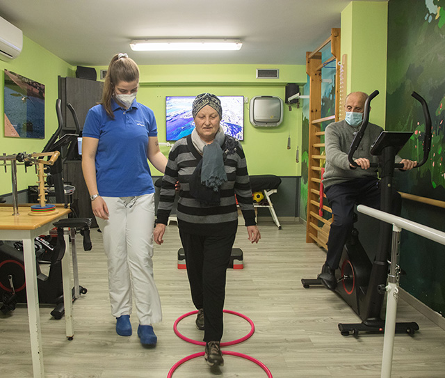 Imagen de la sala de rehabilitación de la Residencia Rodríguez de Andoin V, de paredes verdes y con iluminación artifical. Una mujer mayor esta realizando sus ejercicios, mientras una monitora vestida con polo azul y pantalones blancos le sujeta del brazo para ayudarla. Detrás de ellas, un señor mayor en una bici estática las observa.